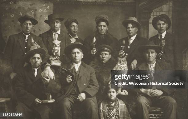 Posed studio group portrait in front of a painted backdrop of Winnebago Indian men and one young woman, Black River Falls, Wisconsin, 1910. Each of...