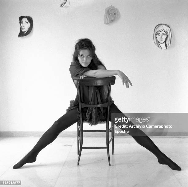 Italian actress Claudia Cardinale straddling a chair, wearing black tights and a black scarf in an empty room with drawn faces hanging on the back...