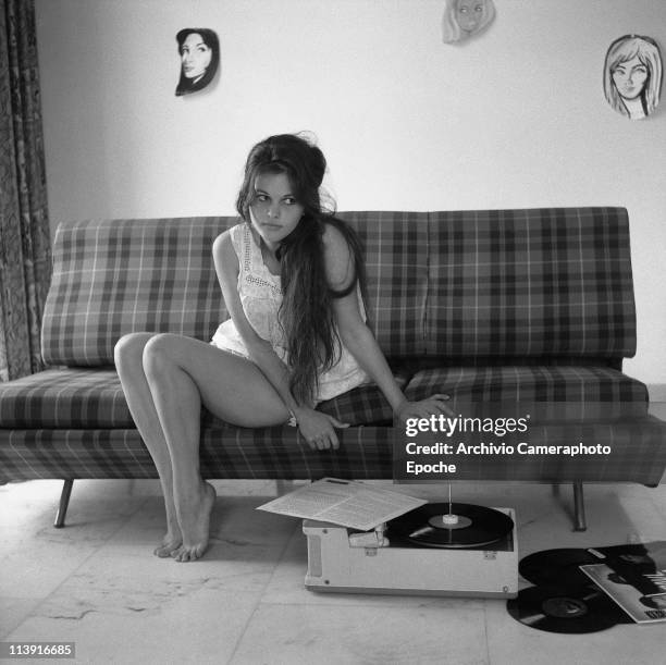 Italian actress Claudia Cardinale sitting on a plaid sofa listening to Ella Fitzgerald vynils, with drawn faces hanging on the back wall, Rome 1959.