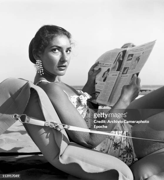 American-born Greek opera singer, Maria Callas lying on an air mattress on Venice Lido beach, wearing a floral swimsuit and dangling earrings,...