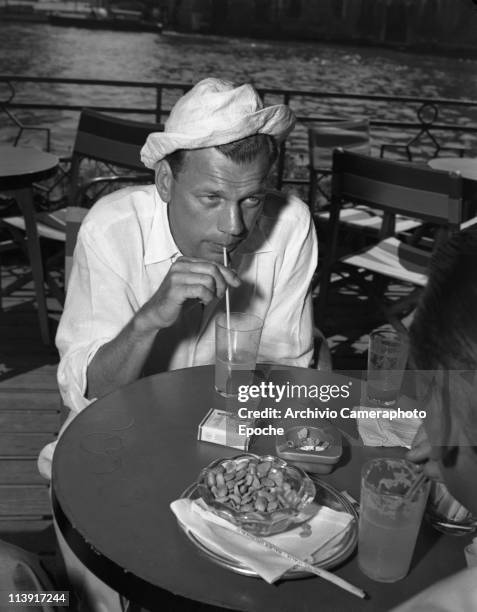 American actor Joseph Cotten, wearing a fabric hat and a linen shirt, sipping a lemonade with a straw sitting at the Bauer Grunwald Terrace. On the...