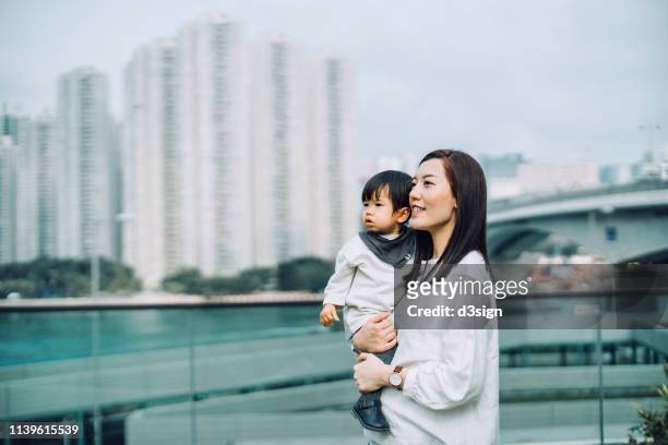 young asian mother carrying daughter standing on urban balcony overlooking city view against urban cityscape - über etwas schauen stock-fotos und bilder