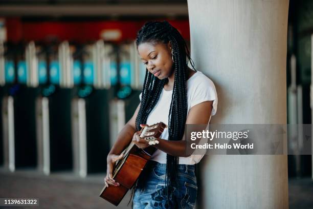 beautiful braided hair young woman playing guitar whilst leaning against a pillar - busker stockfoto's en -beelden