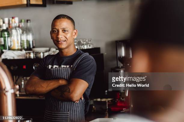 Apron wearing barman or chef looking ointo camera with folded arms and the out of focus head of customer in foreground