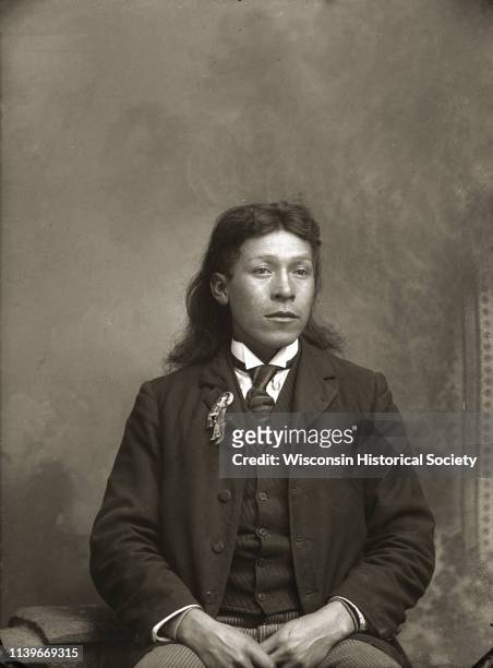 Waist-up studio portrait in front of a painted backdrop of a Ho-Chunk man with long hair posing sitting, Black River Falls, Wisconsin, 1900. He is...