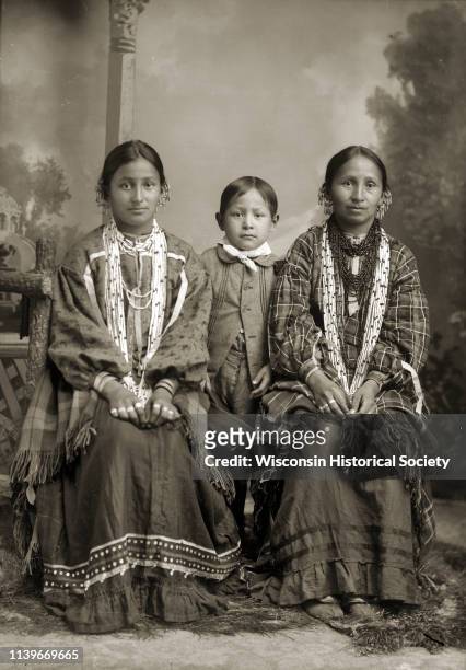 Full-length studio portrait of two Ho-Chunk women posing sitting in front of a painted backdrop near a prop wooden fence, Black River Falls,...