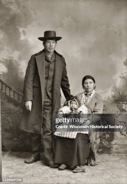 Full-length studio portrait of a Ho-Chunk man posing standing, and a Ho-Chunk woman posing sitting in front of a painted backdrop, Black River Falls,...