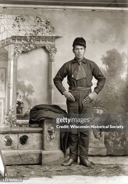 Studio full-length portrait in front of Van Schaick's gazebo backdrop of a Ho-Chunk man, wearing period clothes, standing next to a prop stone wall...