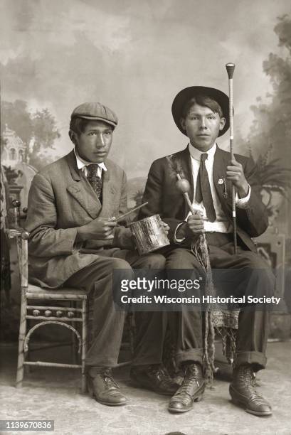 Full-length studio portrait of two Ho-Chunk men posing sitting in chairs in front of a painted backdrop, Black River Falls, Wisconsin, 1920. Sam...