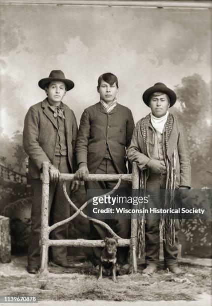 Studio portrait in front of a painted backdrop of three Ho-Chunk men posing standing behind a prop wooden fence, Black River Falls, Wisconsin, 1890....