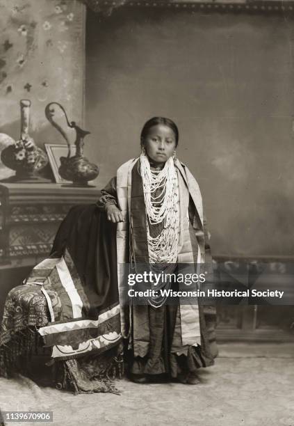 Full-length studio portrait in front of a painted backdrop of a Ho-Chunk girl posing and standing with her arm resting on a chair with a floral...