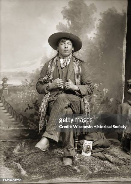Studio portrait in front of a painted backdrop of an unidentified Ho-Chunk man with long hair posing sitting with his legs crossed, Black River...