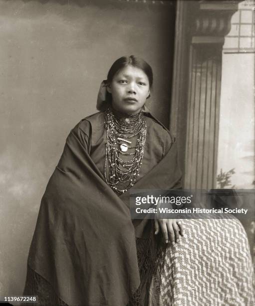 Three-quarter length studio portrait of a Ho-Chunk woman posing standing in front of a painted backdrop with her left hand on a draped chair, Black...