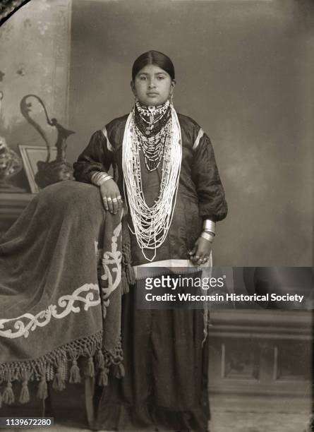 Studio portrait of a young Ho-Chunk woman posing standing in front of a painted backdrop, Black River Falls, Wisconsin, 1909. Her right arm is...