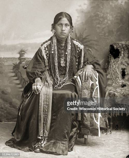 Studio portrait of Ho-Chunk woman, Mabel White Blackhawk St Cyr sitting in front of a painted backdrop with her left arm resting on a silk applique...