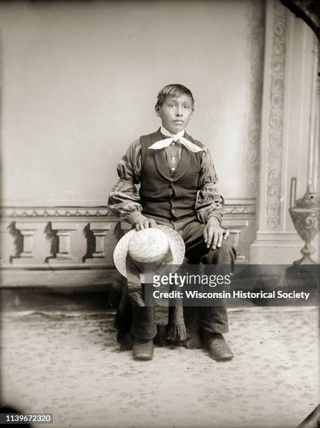 Full-length studio portrait in front of a painted backdrop of an unidentified boy holding a hat with a wide ribbon band on his knee, Black River...