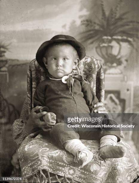 Studio portrait in front of a painted backdrop of a Ho-Chunk infant boy posing sitting in a chair draped with a cloth, Black River Falls, Wisconsin,...