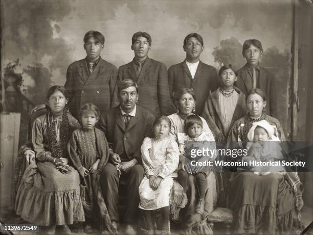 Studio portrait of a large Ho-Chunk group posing in front of a painted backdrop, including five men, three women, three girls, and two smaller...