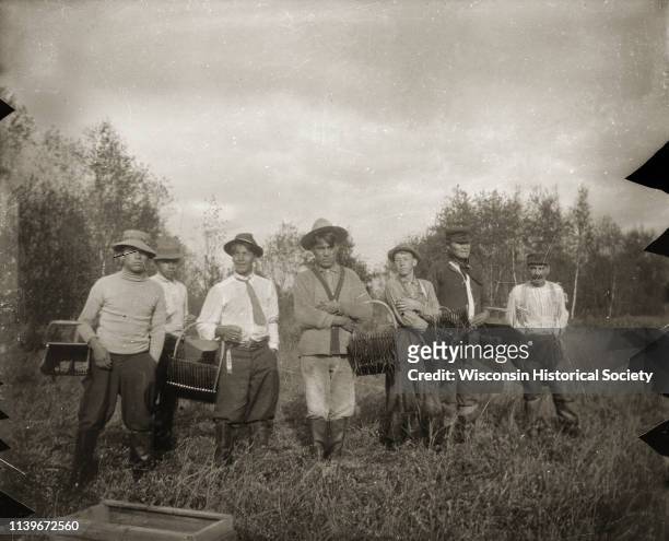 Outdoor group portrait of seven Ho-Chunk men standing outdoors in a cranberry bog, They are holding the hand scoops used to harvest cranberries,...