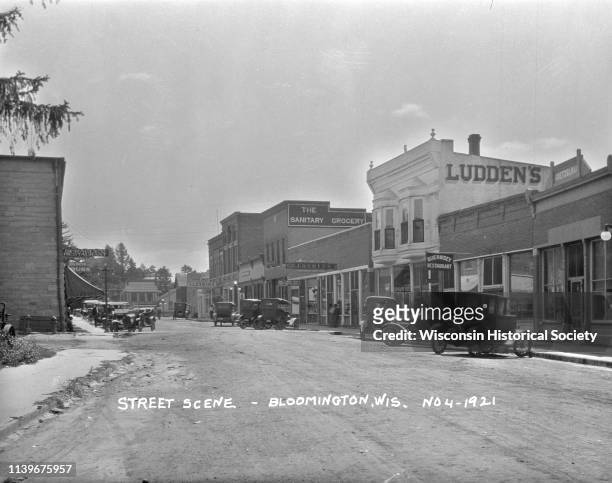 Cars are parked at the curbs along the shops, Bloomington, Wisconsin, 1921. The visible shop signs are Guernsey Restaurant, Ludden's, Ludden's Store...