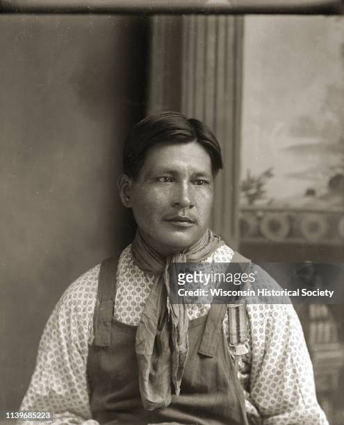 Waist-up studio portrait of a Ho-Chunk man, Henry Snake , sitting and wearing a scarf around his neck in front of a painted backdrop, Black River...