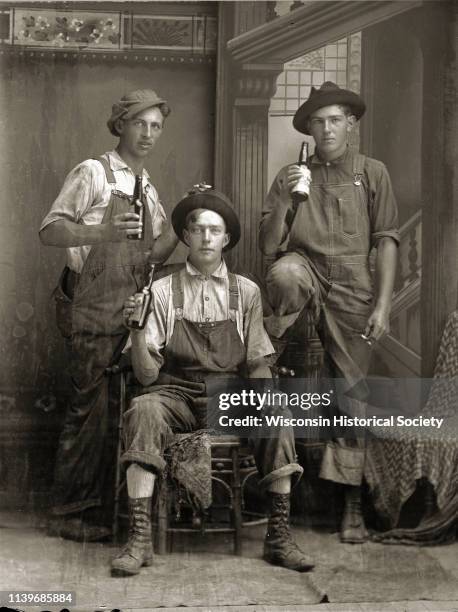 Full-length studio portrait in front of a painted backdrop of three unidentified white men dressed in bib overalls, Black River Falls, Wisconsin,...