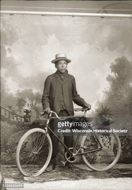 Full-length studio portrait of a young Ho-Chunk man, Jim Carriman, posing standing and holding a bicycle in front of a painted backdrop, Black River...