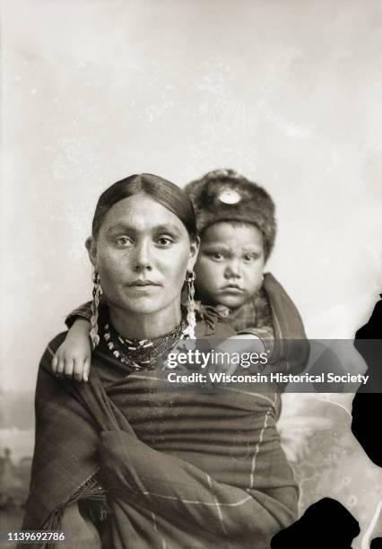 Quarter-length studio portrait of a woman in Ho-Chunk regalia wrapped in a blanket, with a child on her back, Black River Falls, Wisconsin, 1887. The...