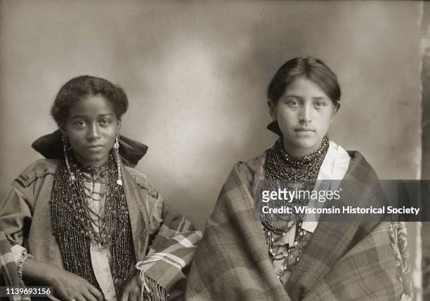 Waist-up studio portrait of two young Ho-Chunk women posing sitting, wearing beaded necklaces and long earrings, and wrapped in shawls, Black River...