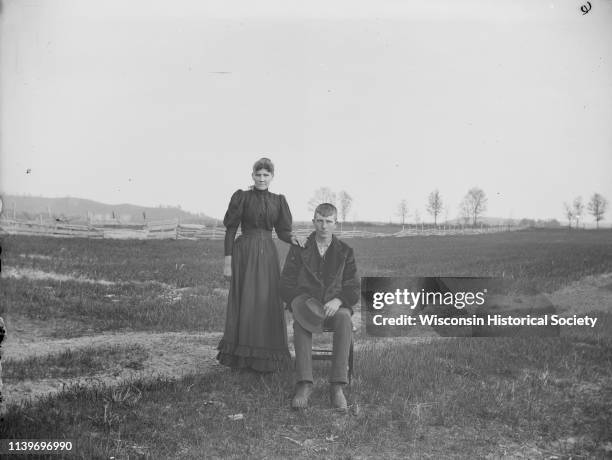 Man sitting on chair, and woman standing with hand on his shoulder, in a field with a wooden fence in the background, Black River Falls, Wisconsin,...