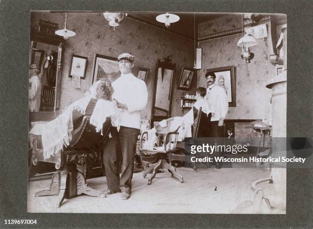 The interior of a barbershop with customers in chairs and barbers at work, Black River Falls, Wisconsin, 1895. Numerous mirrors, pictures, and lamps...