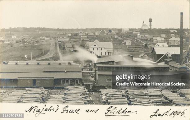 View of a sawmill and the distant town in the background, Glidden, Wisconsin, 1906. Stacks of logs are in the lumberyard. The postcard bears a New...