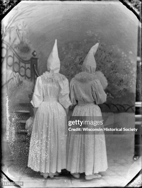 Studio portrait of two women wearing white pointed hats and long, white dresses with fluffy collars, and broad white belts, Black River Falls,...