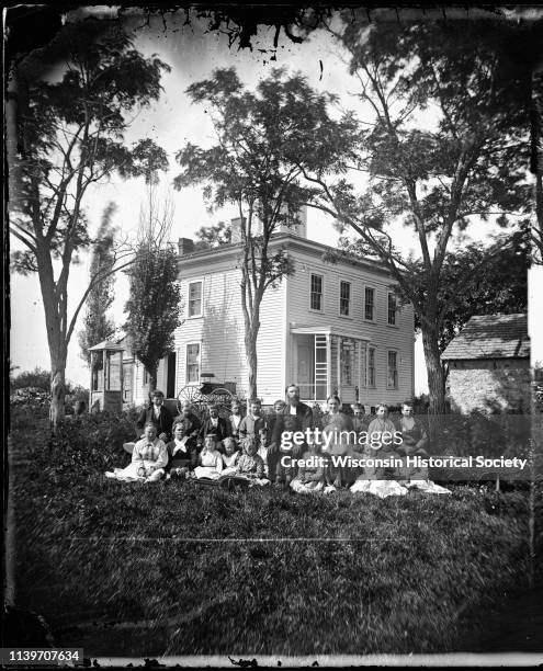 Twenty-one children are seated on wooden benches and the ground in front of a square frame house with a hip roof and cupola, Clinton, Wisconsin,...