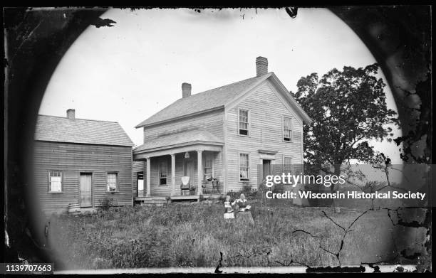 Family plays croquet in the yard of a side gable frame house with a porch, Wisconsin, 1874. A bird cage hangs from the porch and a young girl...