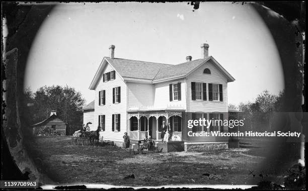 Seven people are grouped around an impressive two-story home with a fan window at the attic gable and a porch with carpenters lace and latticework,...