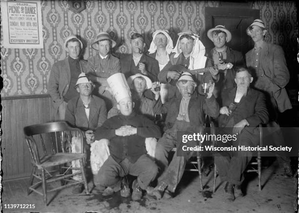 Group of a dozen men mugging for the camera, Black River Falls, Wisconsin, May 1913. Poster on the wall advertises a 'Grand Picnic and Dance in...