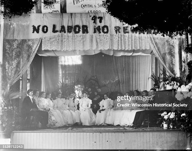 Two young men and ten young women in white dresses sitting in a semi circle on the stage of the Black River Falls Opera House, Black River Falls,...