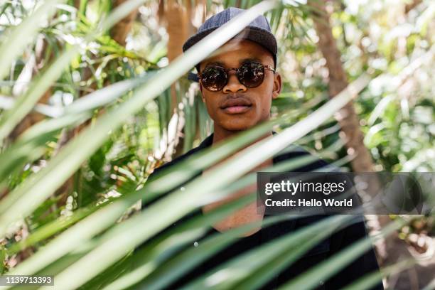 handsome young man wearing sunglasses and peak cap seen through palm frond in foreground - sunglasses disguise stock pictures, royalty-free photos & images