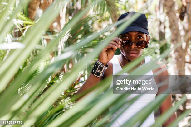 handsome young man removing sunglasses and looking into camera seen through palm frond in foreground - sunglasses disguise bildbanksfoton och bilder