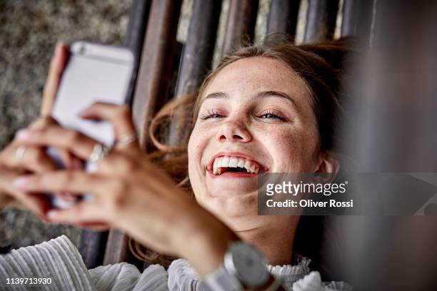 laughing young woman lying on a bench using cell phone - women lying imagens e fotografias de stock