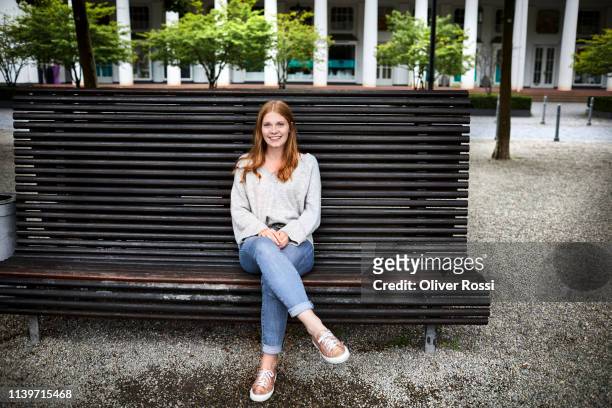 smiling young woman sitting on a bench - sitzen stock-fotos und bilder