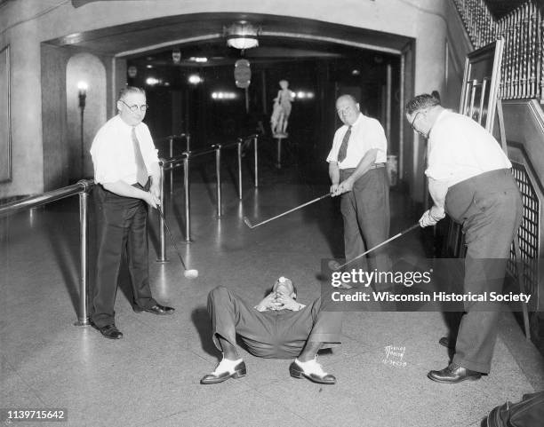 Men posing during a golf stunt, as they try to hit a golf ball off a man's nose while he is lying on his back on the floor in the lobby of the...