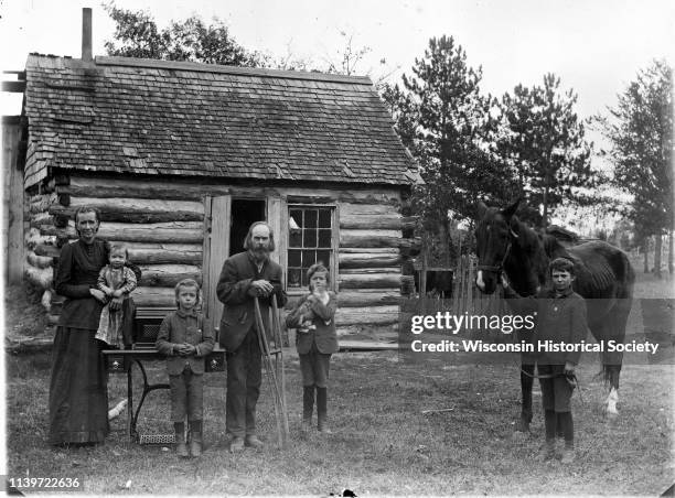 Farm family posing in front of a log home, Black River Falls, Wisconsin, 1900. There is a man on crutches in the center, a woman is holding a small...