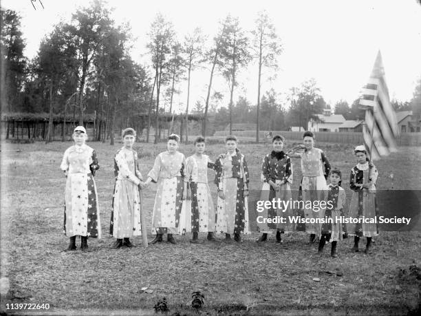 Nine girls are posing standing and wearing patriotic costumes, Black River Falls, Wisconsin, 1904. One girl is holding a flag of the United States....