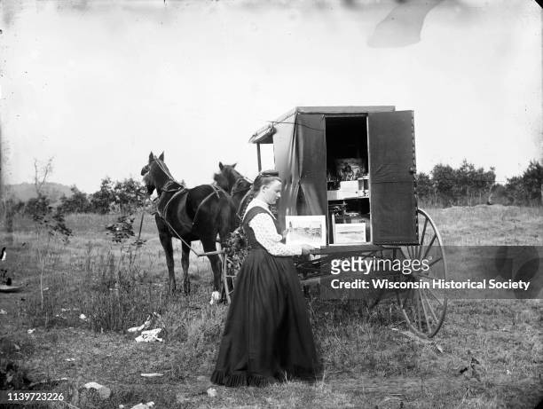 Woman, possibly the daughter of CR Monroe, holding up an image of Black River Falls Main Street at the back of a traveling photographer's wagon,...