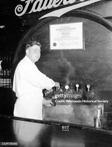 Apron-clad 'Moon' Molinaro, Fauerbach's bartender, fills a stein of Fauerbach beer from the tap, with Arthur Towell's University of Wisconsin diploma...