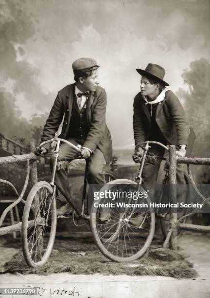 Full-length studio portrait of two young Ho-Chunk men posing sitting on bicycles near a prop wooden fence in front of a painted backdrop, Black River...