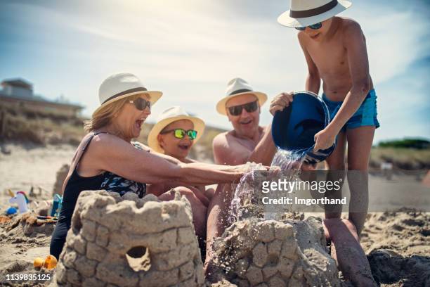 abuelos y nietos jugando juntos en la arena en la playa - castillo de arena fotografías e imágenes de stock