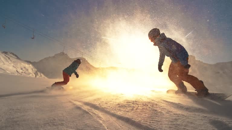 Wide speed ramp tracking shot of two snowboard riders riding towards the setting sun with snow particles flying in the air. Shot in Austria.